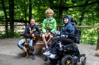 Junger Elektrorollstuhlfahrer und zwei Kletterer auf einer Bank im Wald