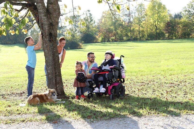 Familie auf einer Wiese mit Baum rollstuhlfahrendes Kind