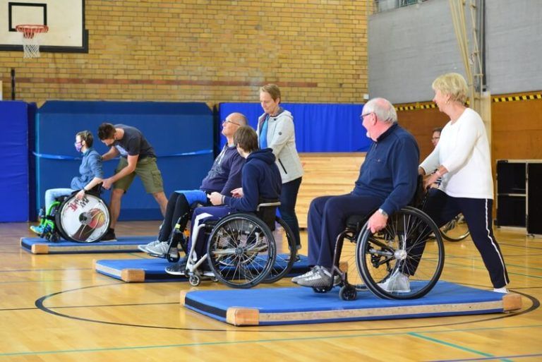 Rollstuhlfahrer mit Begleitern in einer Sporthalle auf Matten beim Training