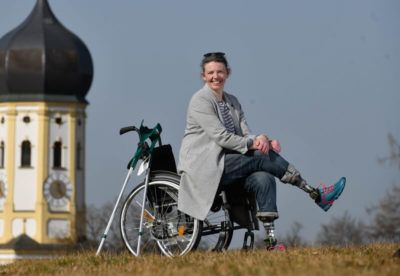 Frau mit Prothesen sitzt im Rollstuhl auf einer Wiese vor einer Kirche