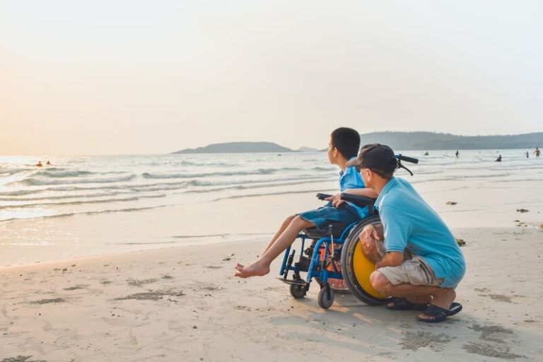 Kind im Rollstuhl mit Vater am Strand, beide schauen aufs Meer hinaus