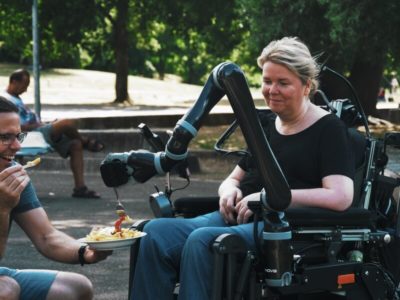 Frau im Rollstuhl, die mithilfe eines Roboterarms Pommes isst