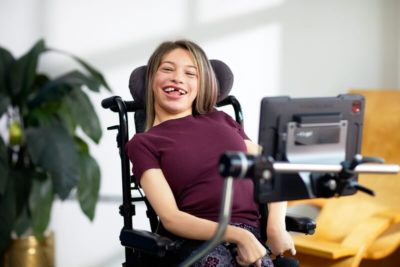 Junge Frau mit Behinderung schaut lachend auf ihr iPad, das sie mit den Augen bedienen kann