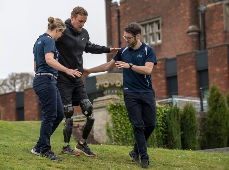 Mann mit zwei Beinprothesen übt das Gehen mit Hilfe von zwei Therapeuten
