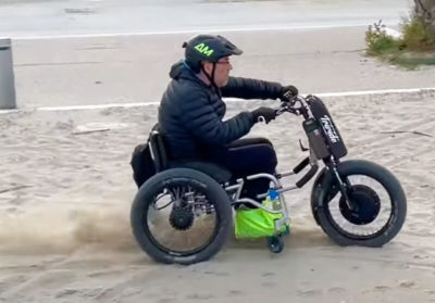 Man sieht einen Rollstuhlfahrer mit Helm und Zuggerät bei einer rasanten Fahrt auf Sand