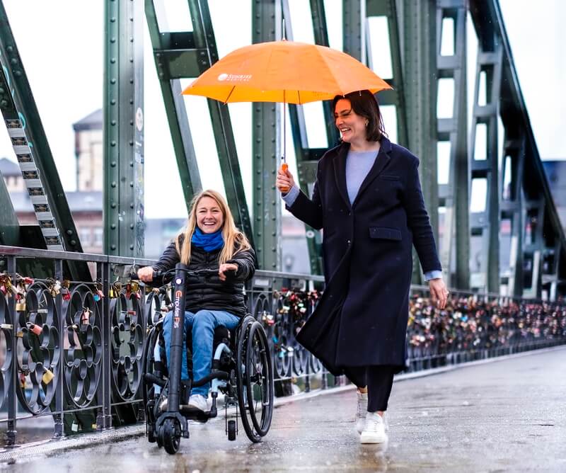 Frau im Rollstuhl mit Zuggerät auf einer Brücke. Es regnet, eine andere Frau hält einen Schirm, beide lächeln.