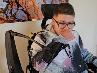 Junger Mann im Rollstuhl kratzt sich dank einer Armunterstützung selbstständig an der Nase