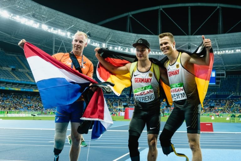 Drei Paralympicsläufer mit Landesflagge über den Schultern im Stadion