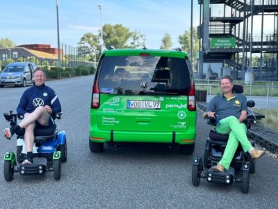 Ein Auto von hinten, daneben sitzen rechts und links zwei Männer in einem Rollstuhl.