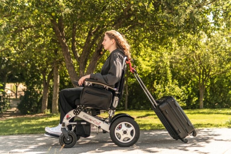 Rollstuhlfahrerin in einem Elektrorollstuhl im Park, die einen Koffer hinter sich herzieht.