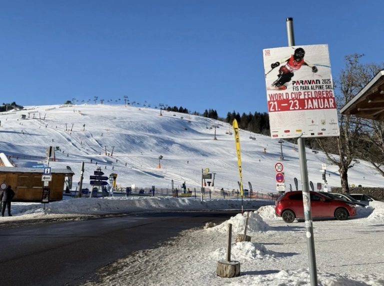 Man sieht eine verschneite Landschaft in den Bergen mit Skipiste, auf einem Plakat im Vordergrund wird eine Weltcupveranstaltung für Menschen mit Behinderung angekündigt