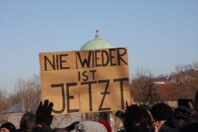 Man sieht ein Pappschild, das auf einer Demo hochgehalten wird, darauf steht "Nie wieder ist jetzt".