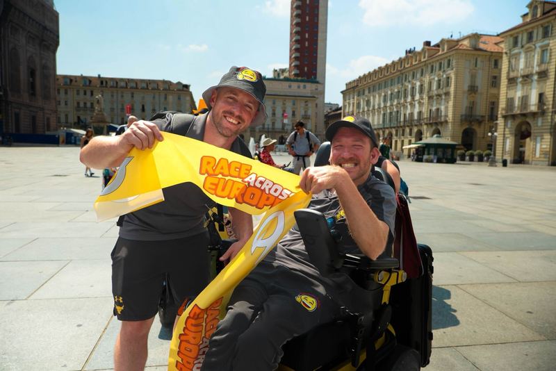 Zwei strahlende Männer feiern auf einem großen Platz in Turin das Ziel des „Race Across Europe“. Einer sitzt im Elektrorollstuhl, der andere steht daneben. Beide tragen Teamkleidung und halten gemeinsam ein gelbes Banner mit der Aufschrift „Race Across Europe“. Im Hintergrund sind historische Gebäude und ein roter Turm zu sehen.