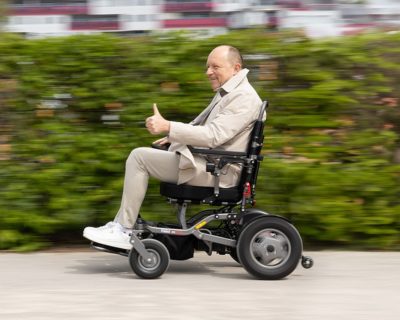 Älterer Herr in einem Elektrorollstuhl, der einen Daumen hebt und offensichtlich mit hoher Geschwindigkeit unterwegs ist, der Hintergrund ist verwischt.