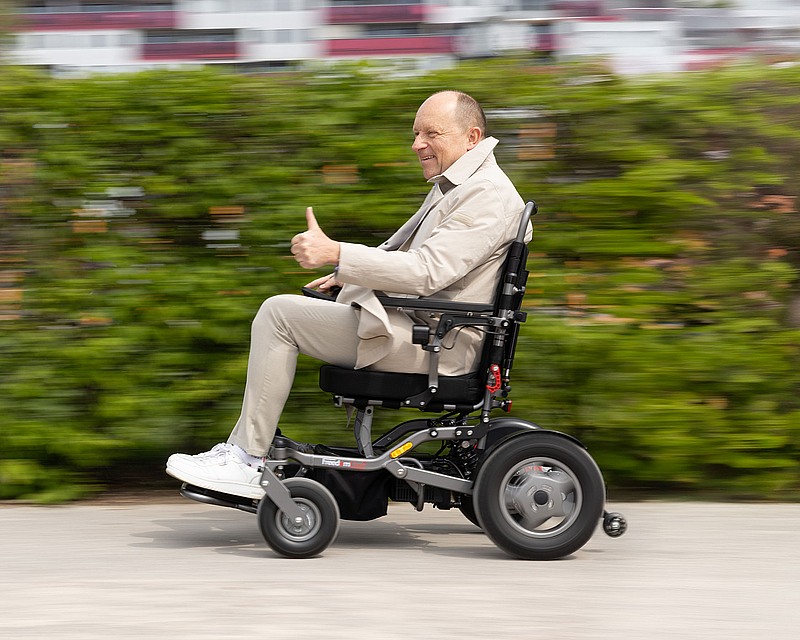 Älterer Herr in einem Elektrorollstuhl, der einen Daumen hebt und offensichtlich mit hoher Geschwindigkeit unterwegs ist, der Hintergrund ist verwischt.